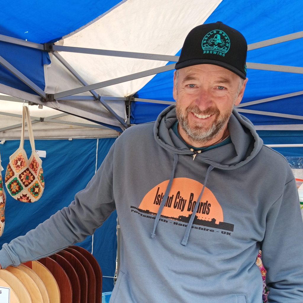 A smiling man wearing a gray hoodie and a black cap stands behind a display of wooden boards labeled 'The Hot Walls Flyer' and 'The Eastney Naturist.'